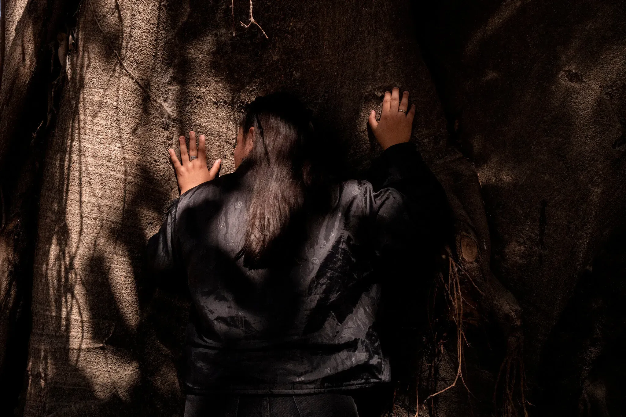 a girl stands against a tree, facing away from the camera