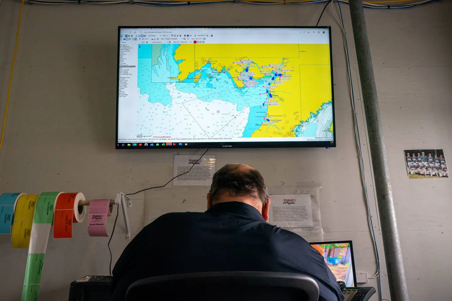A worker sits at a desk in front of a large monitor showing a map of Bristol Bay. Beside him are rolls of multicolored sticker tags and a photo of a children's baseball team. 