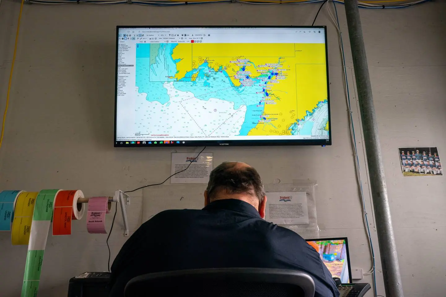 A worker sits at a desk in front of a large monitor showing a map of Bristol Bay. Beside him are rolls of multicolored sticker tags and a photo of a children's baseball team.