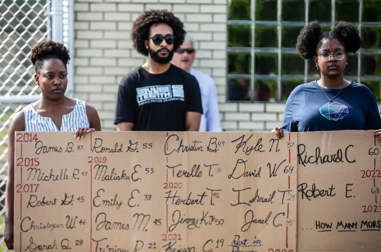 Three people stand holding a cardboard sign with the names of people who died in custody in Pennsylvania jails.
