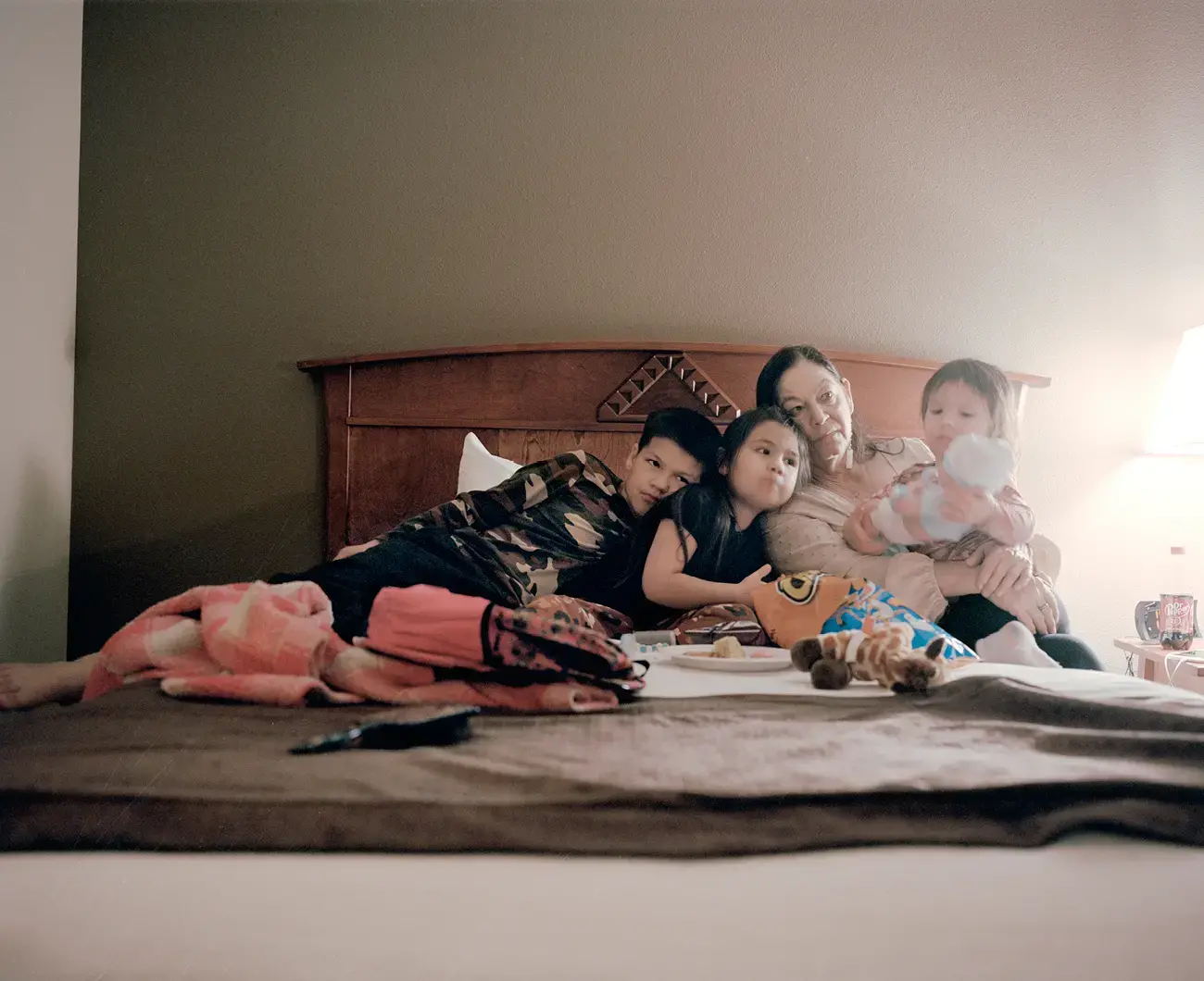 A grandmother sits on a bed with her three young grandchildren. Toys, a backpack, and Cheetos are visible on the bed.