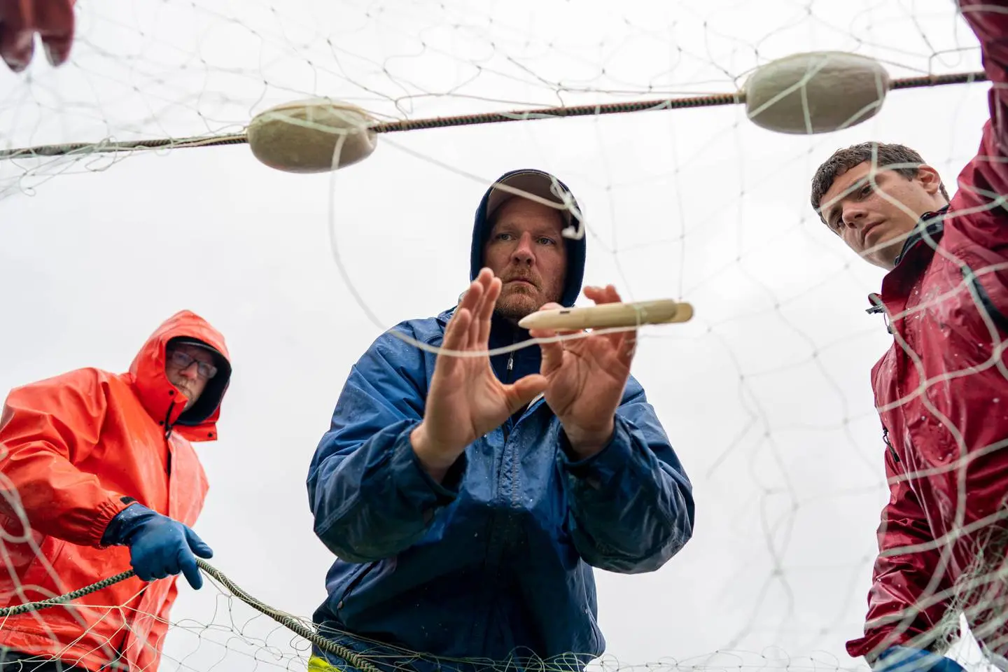 Two fishermen hold a net in place while a third uses a wooden spindle to weave parts of the net together.