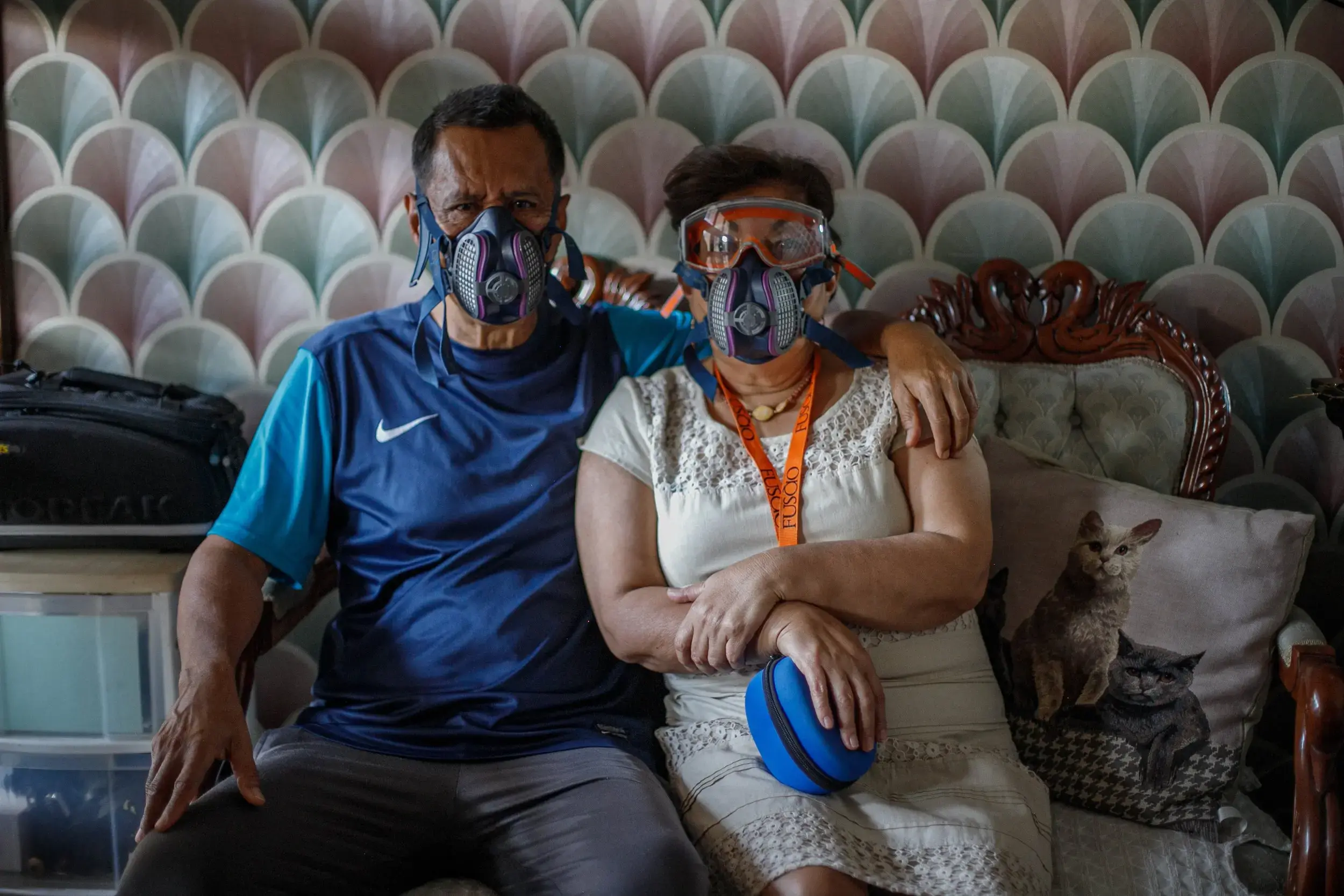 A couple sits next to each other on a couch, wearing protective masks. The women also wears googles. There is a pillow with two cats on the couch.