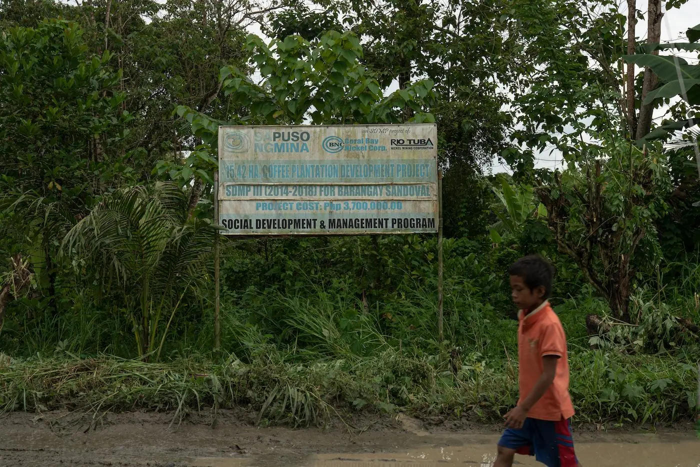 A young boy walks along a muddy road, passing by a worn-out sign about a Rio Tuba Development surrounded by tropical plants by the side of the road. 