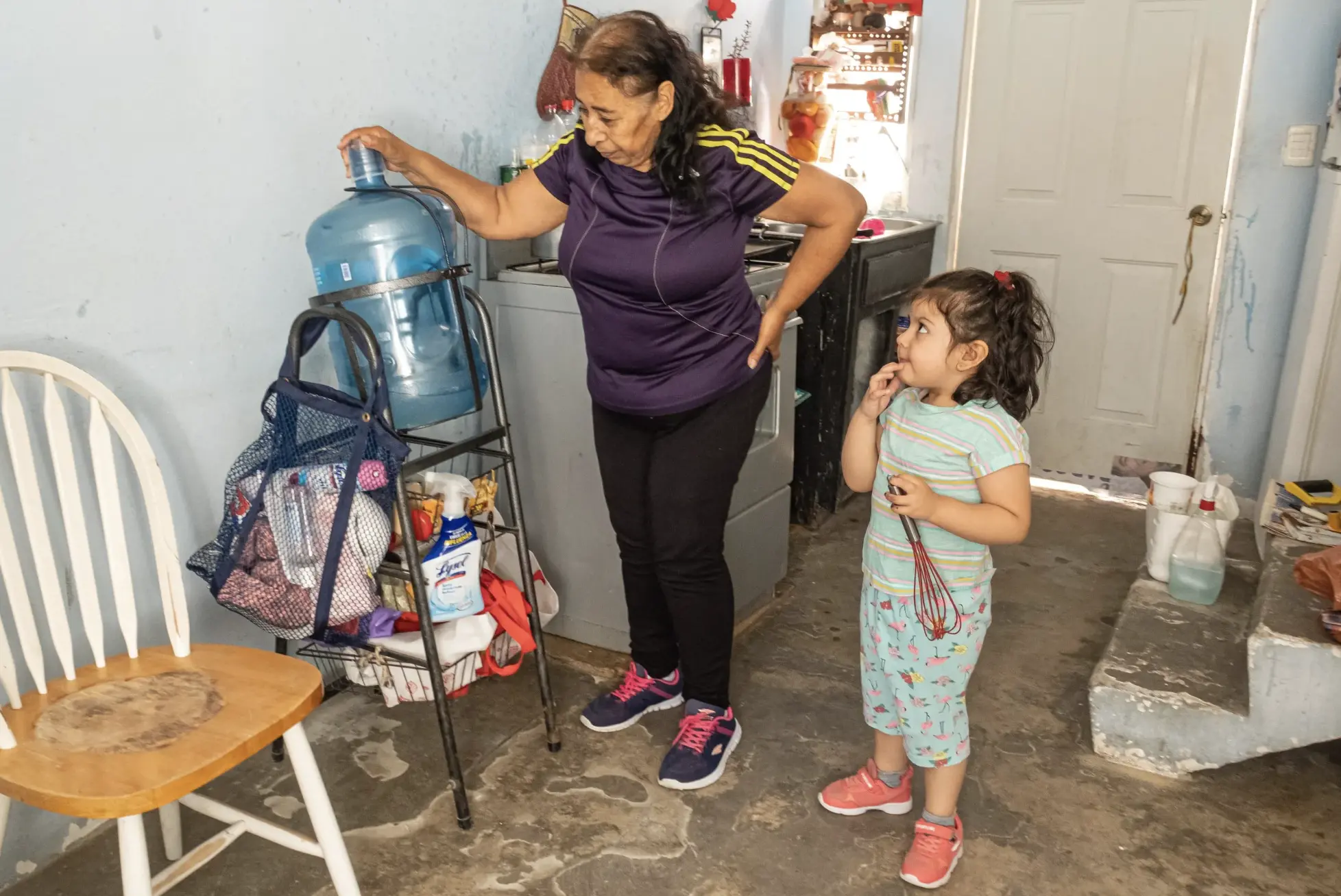 A grandmother stands with granddaughter next to a water jug