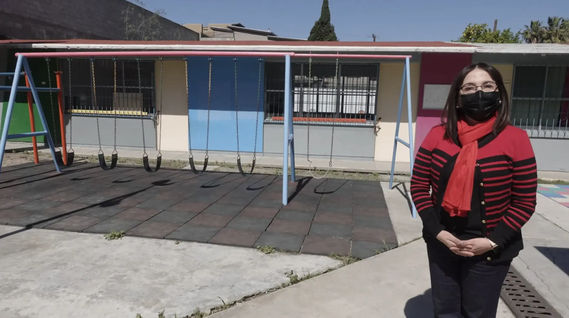 A woman stands masked in a cardigan in front of swings and a preschool