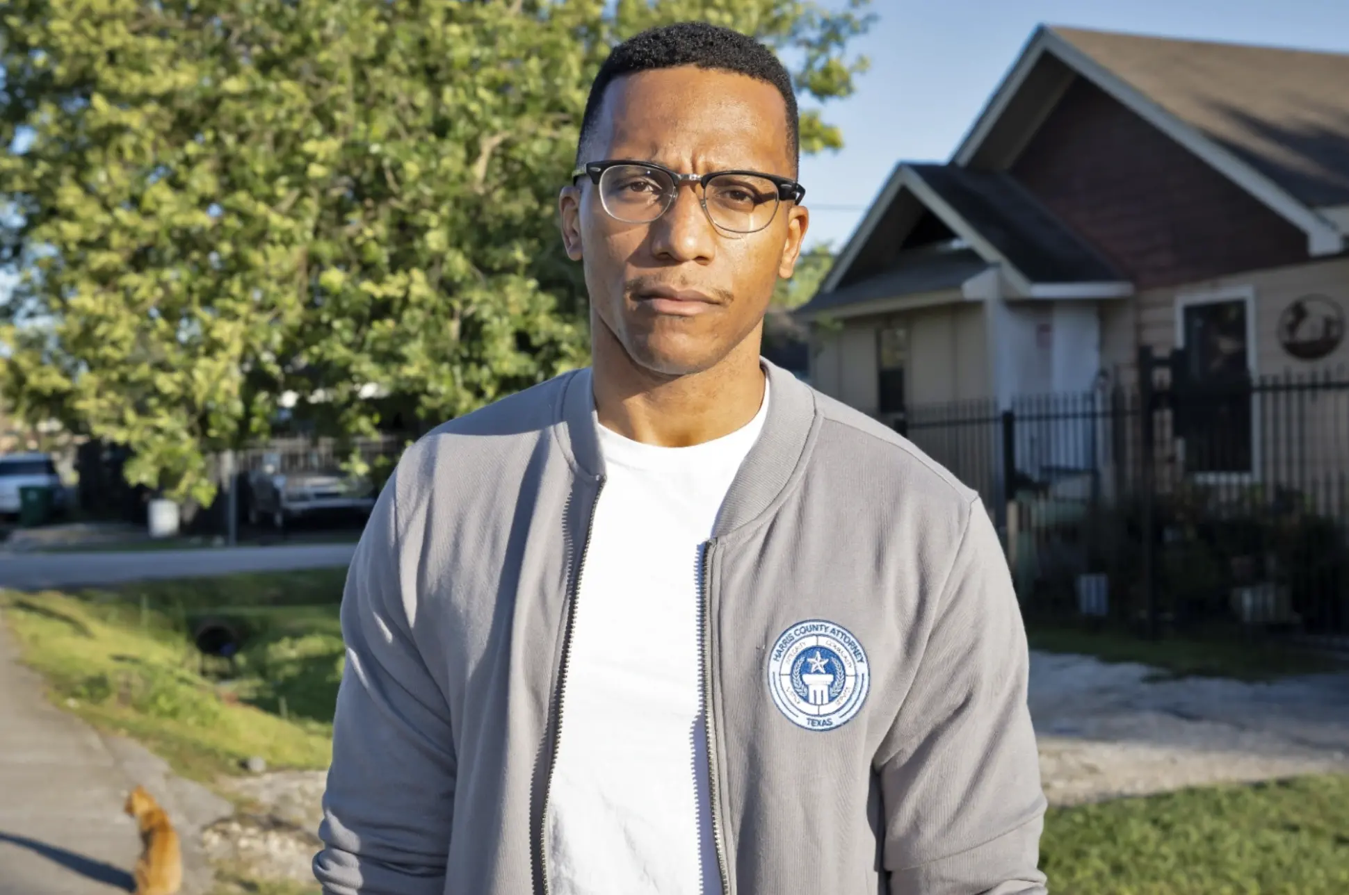 A man with glasses stands in county attorney grey bomber jacket in front of a home and a tree