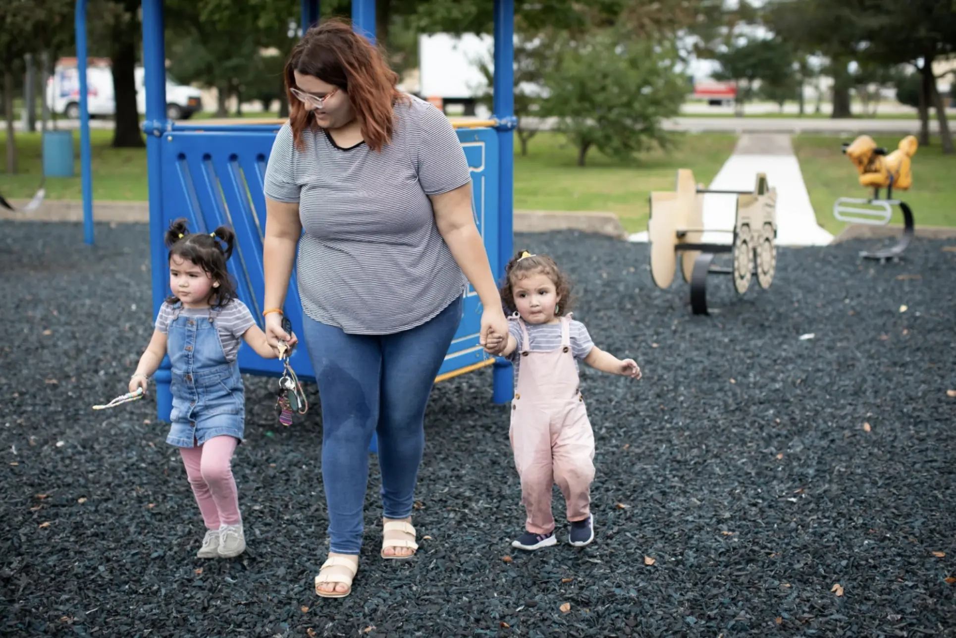 A woman walks across a playground holding her two kids hands