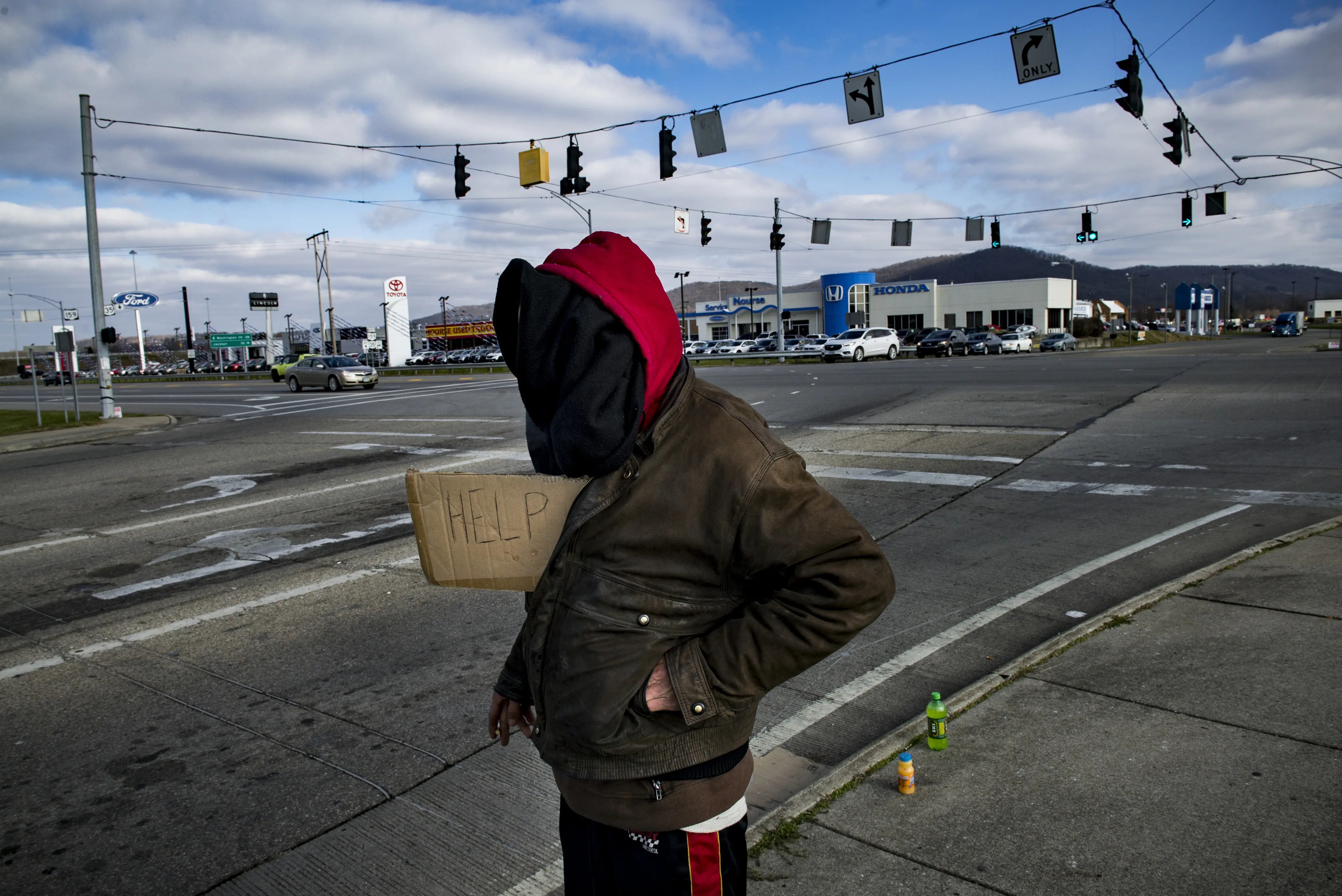 A man, whose face is covered by a black garment, stands at an intersection with a cardboard help sign.