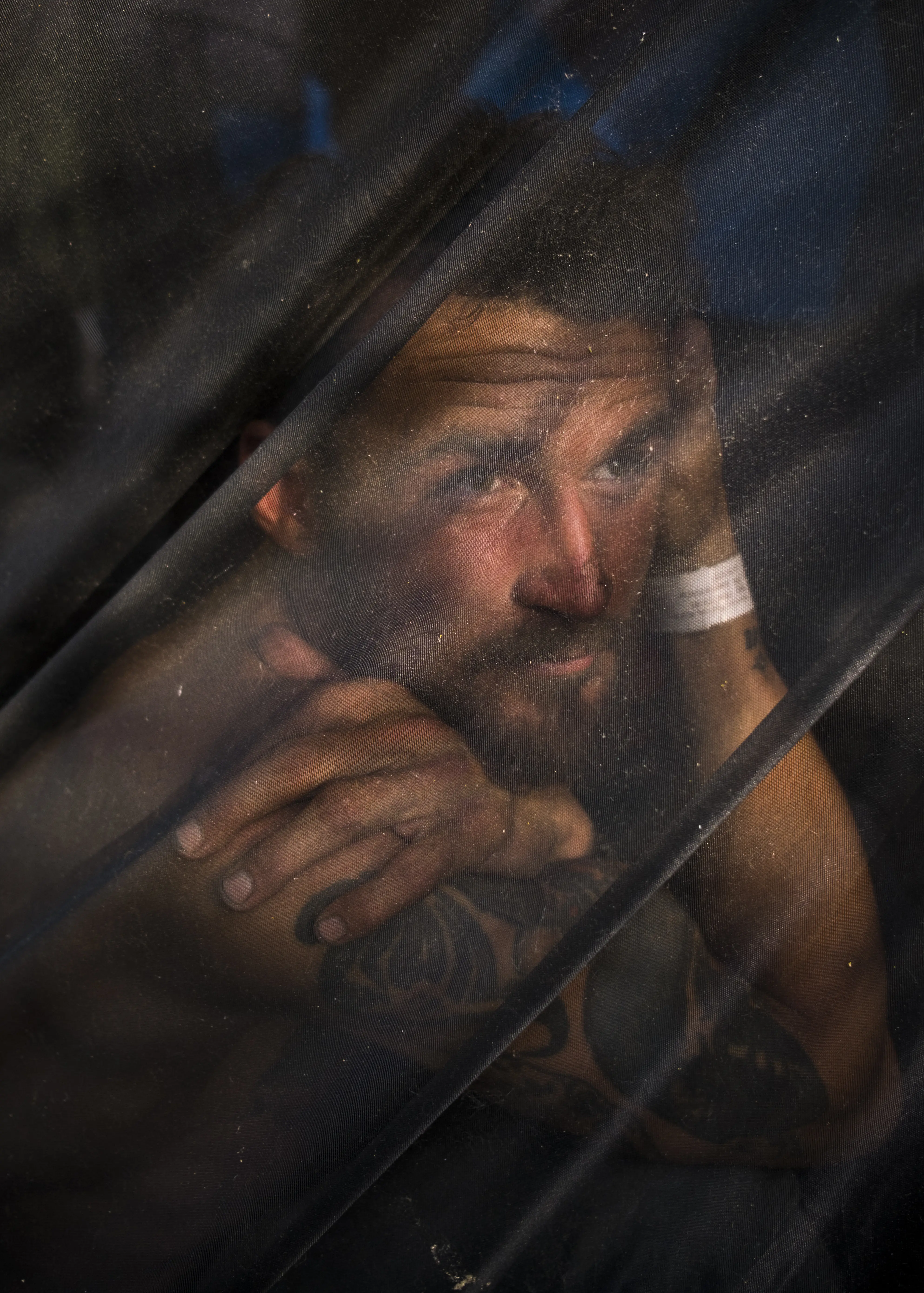 A man, lit dramatically by an out-of-frame source, looks away from the camera through his tent's screen.