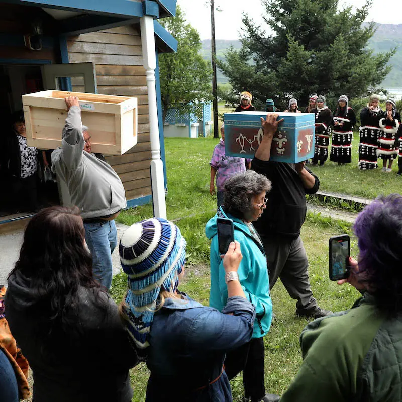 Two men carry boxes on their shoulders while a crowd gathers to watch.