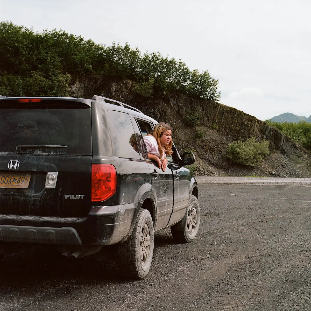 A girl leans out of the back window of an idling, black SUV.