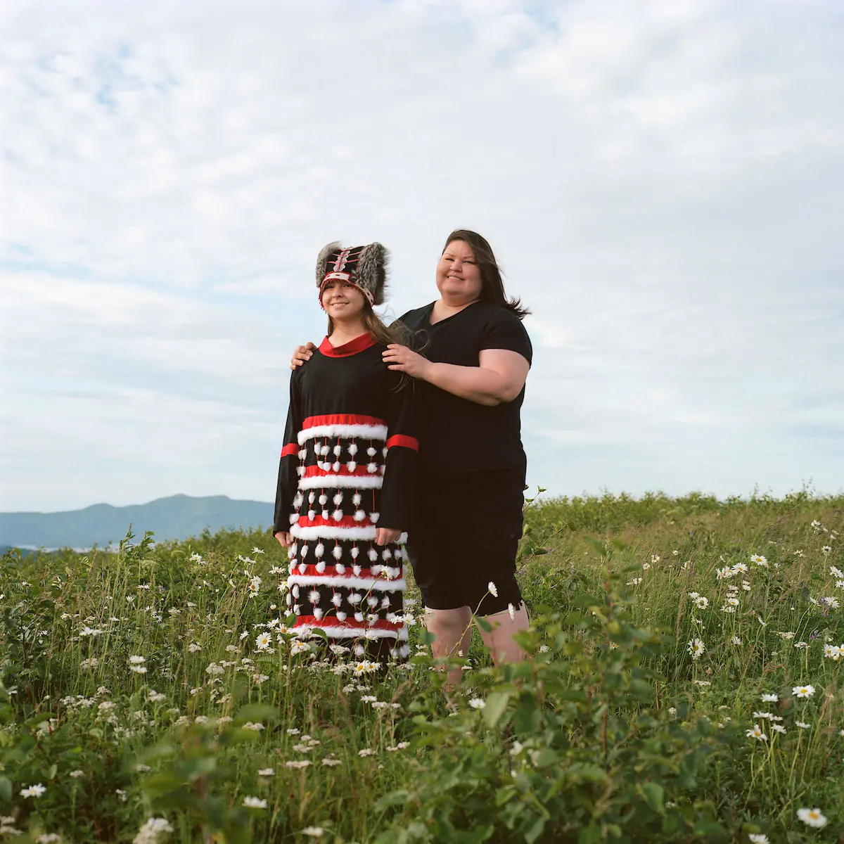 A woman holds a child in regalia by the shoulders. They are in a grassy field.