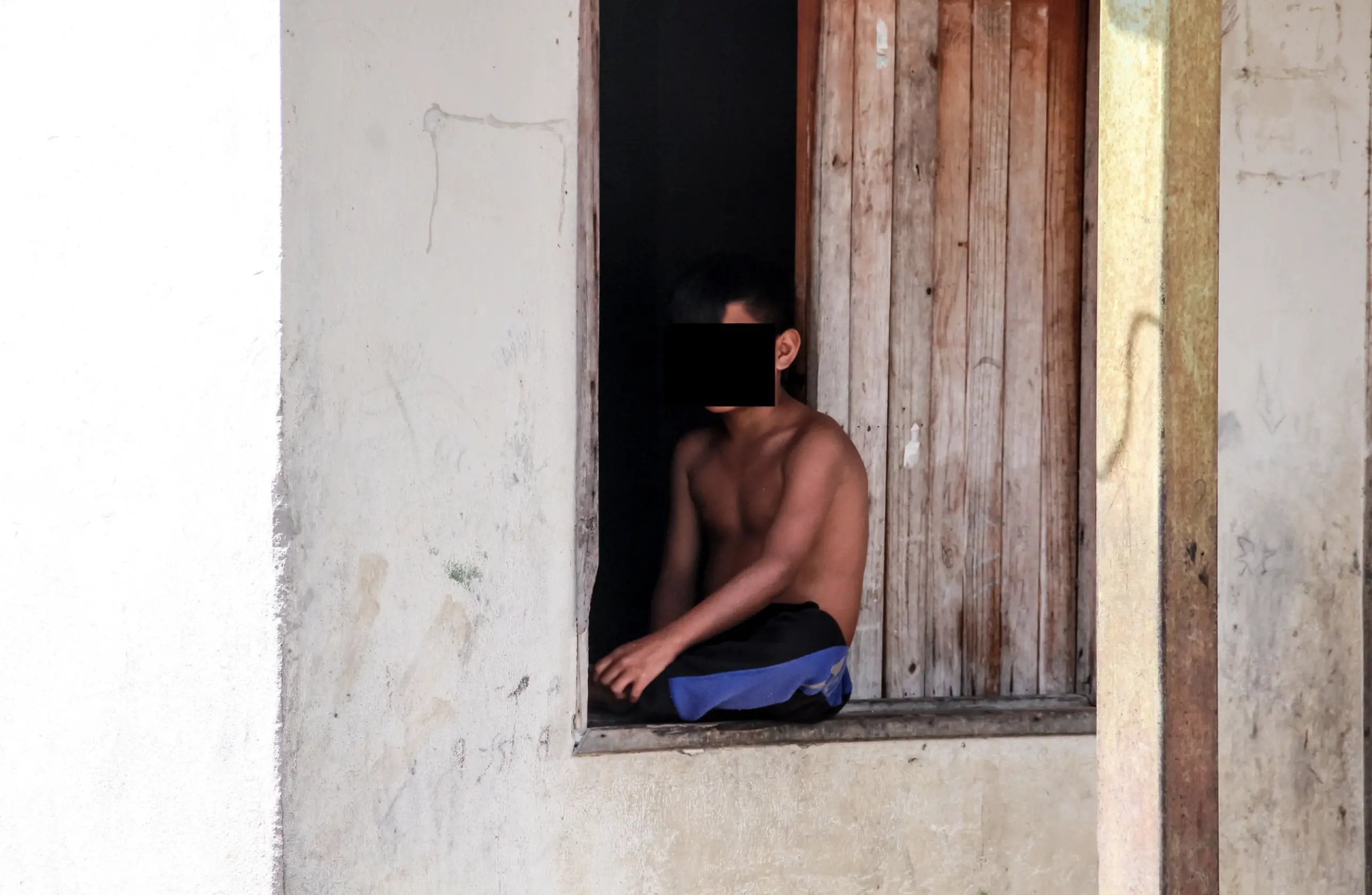 A young boy sits on a windowsill. He is shirtless and wearing shorts. 