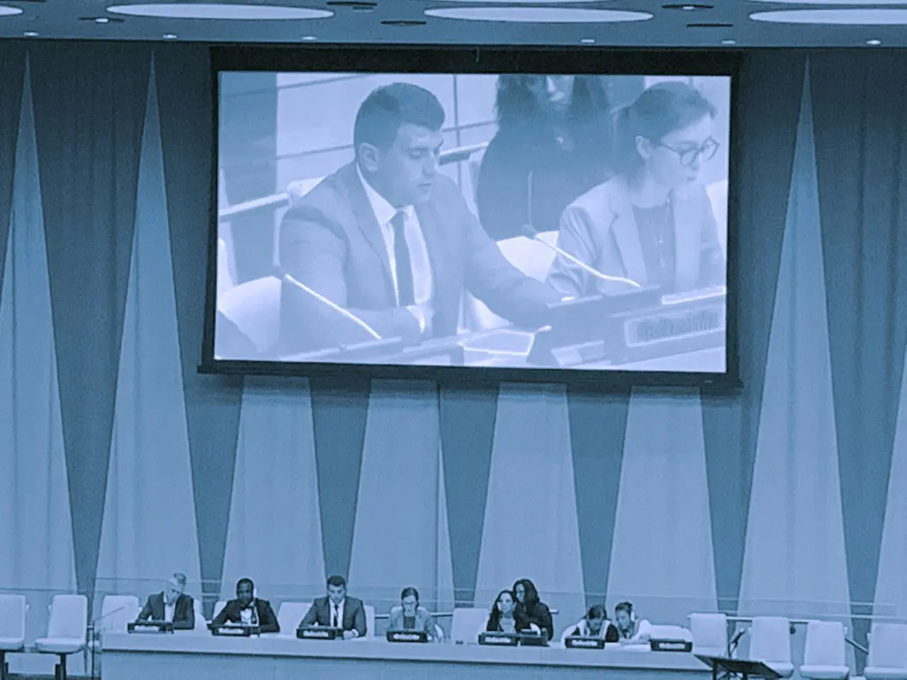 Image of Alen speaking on a projector screen at the UN.