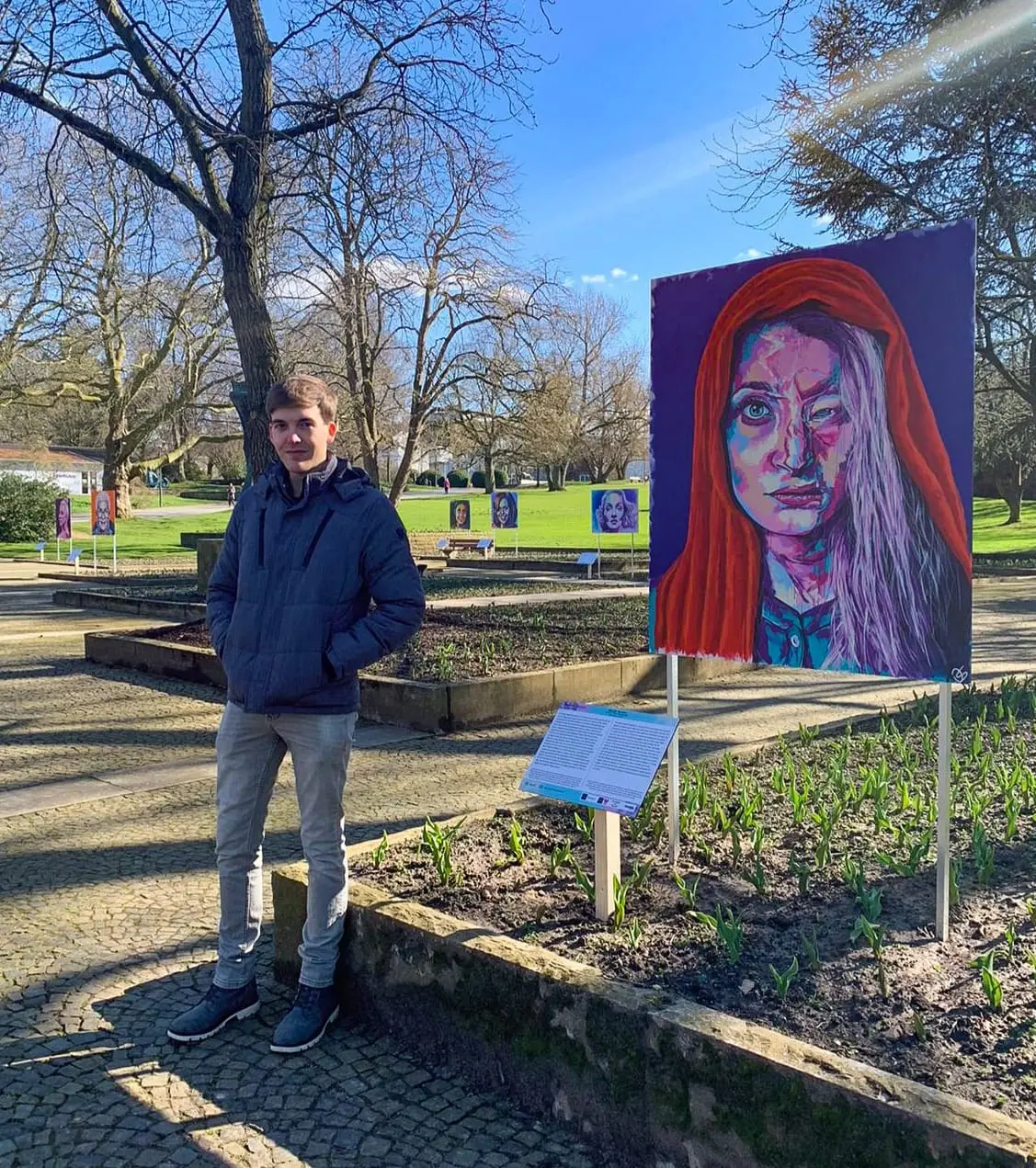 A man stands next to a colorful portrait at an outdoor exhibition.