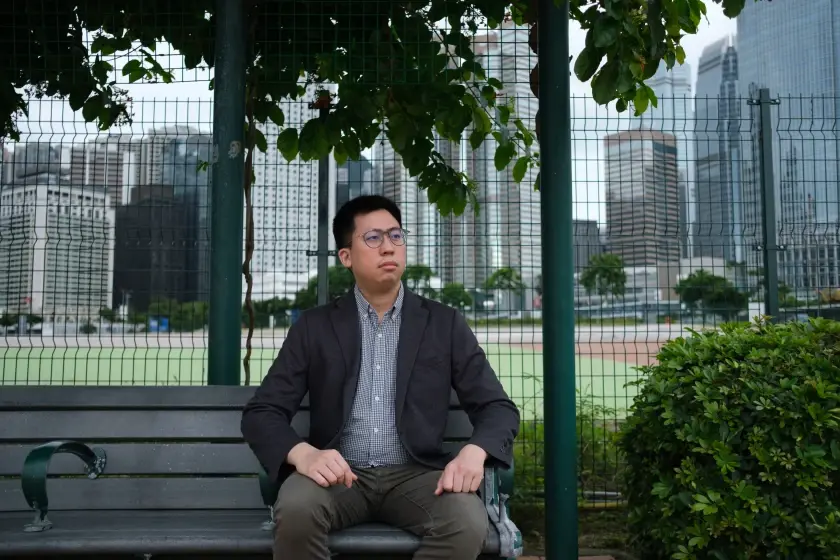 A man on a park bench looks to frame right. A cityscape, viewed through a green mesh fence, is behind him.