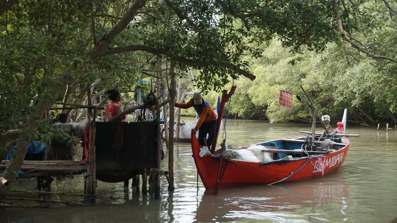 A boat comes in to the river. A woman stands on a bamboo dock while a man orients the boat.
