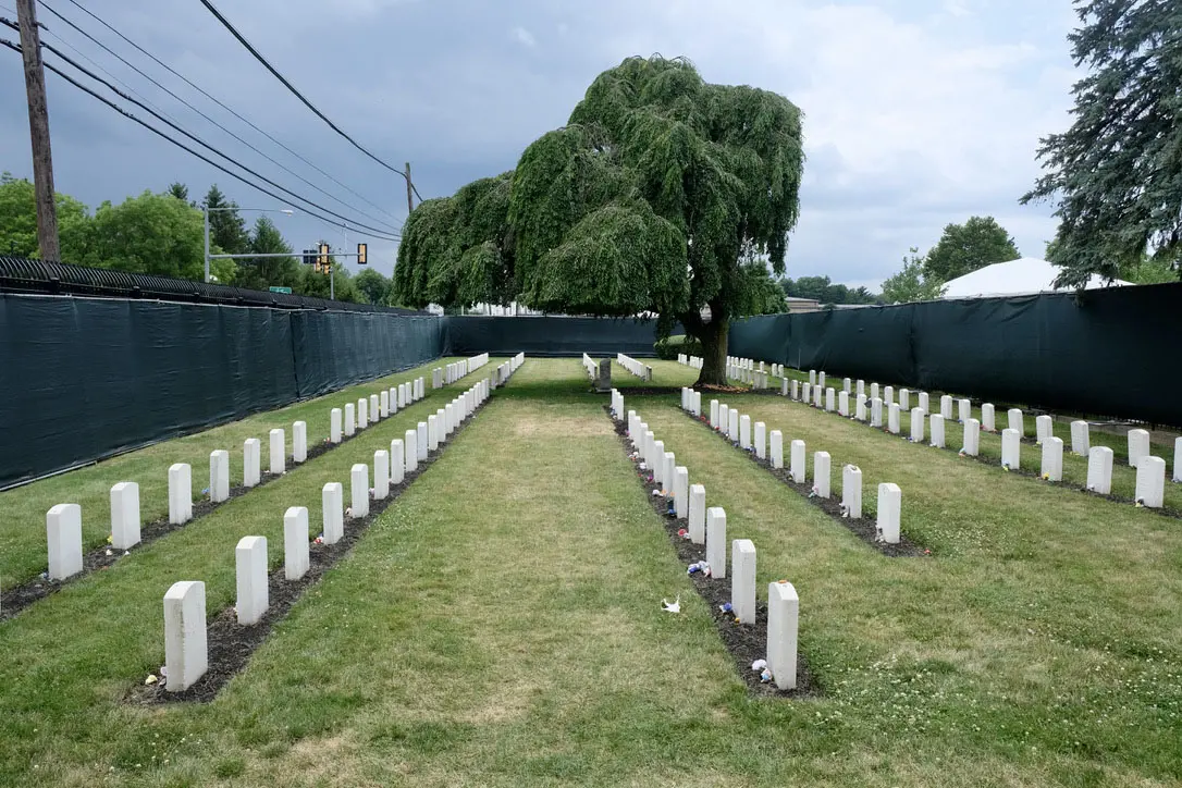 A stooped tree mourns over and shades six uneven rows of headstones; they are fenced in by wire and tarp.