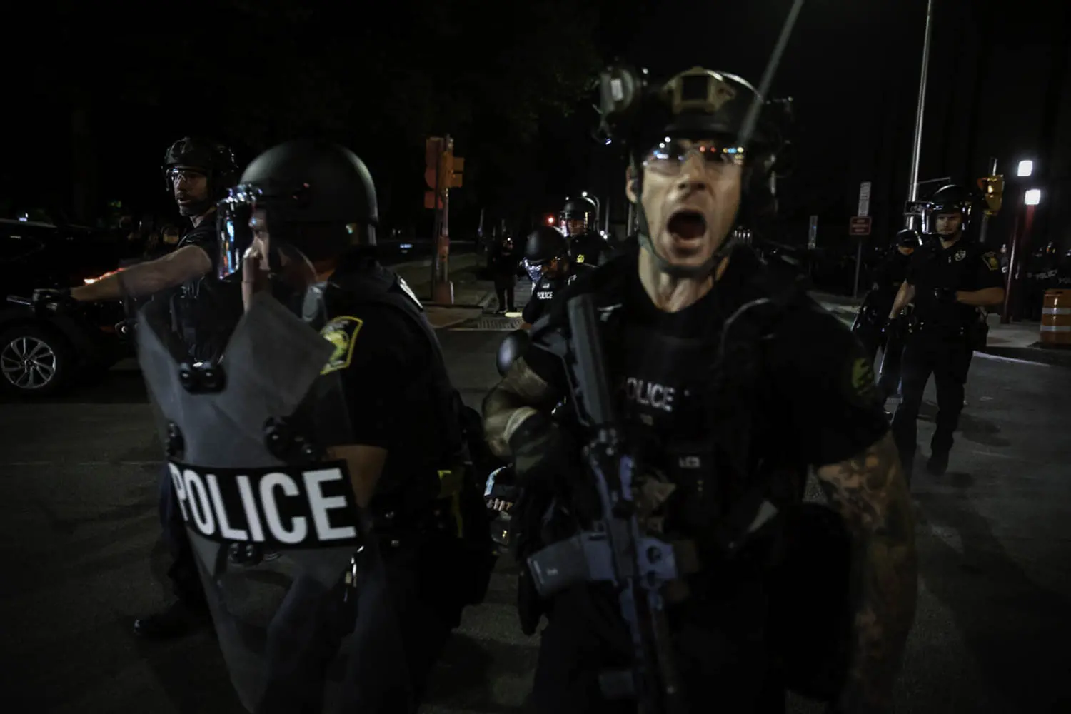 A police officer, carrying an automatic rifle, yells. Other armed officers, carrying shields and wearing helmets, stand around him.