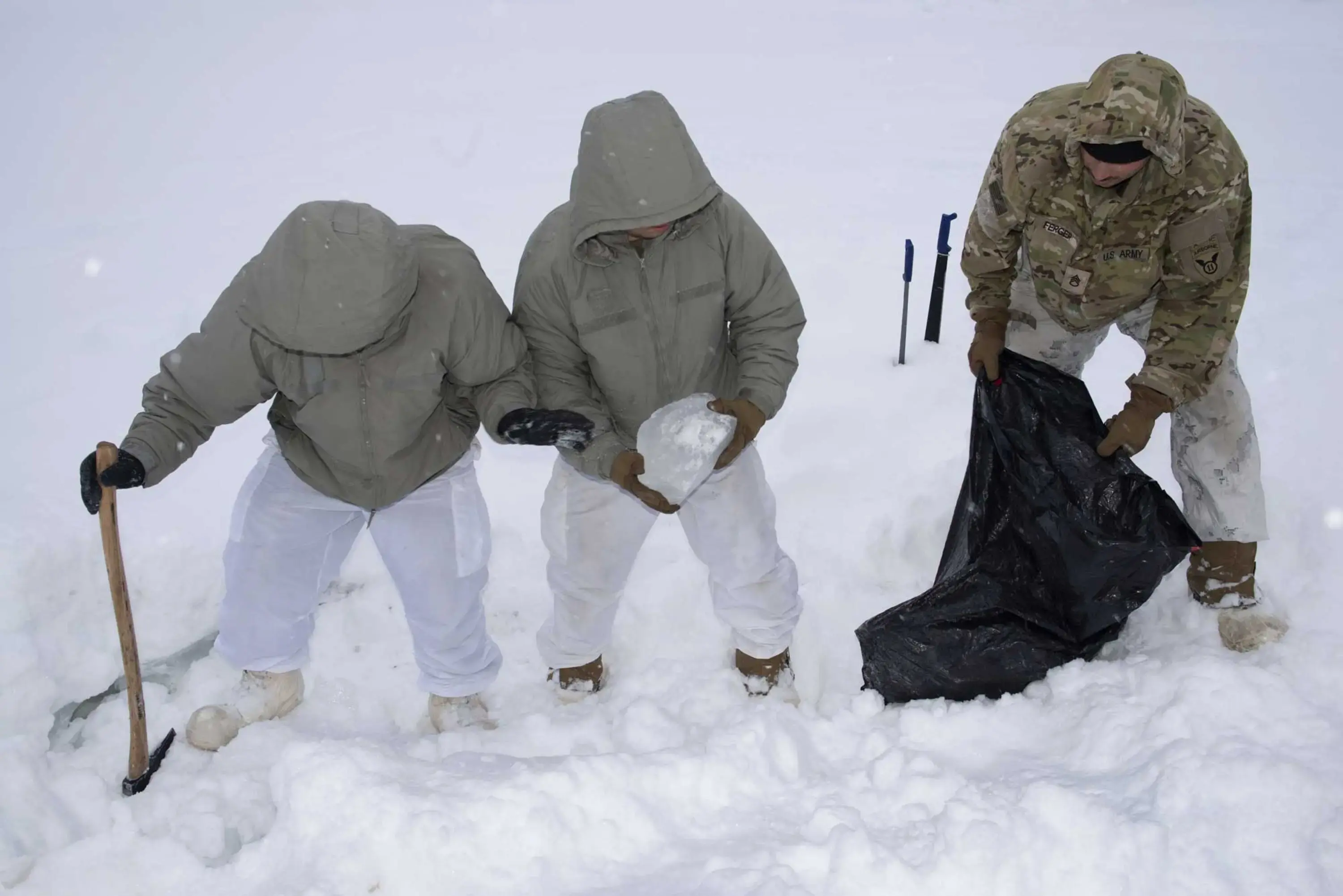 Three soldiers stand in ankle-deep snow: one holds a hatchet, the second holds a chunk of ice and the third is holding a large, black plastic bag.