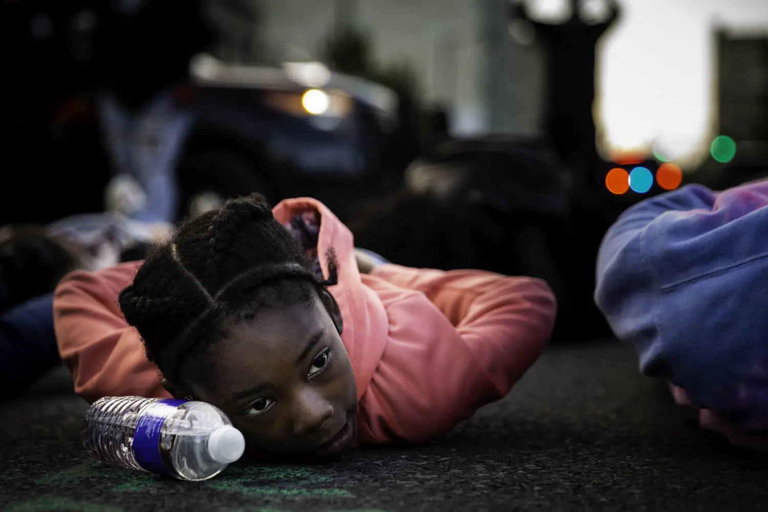 A young girl lies on the ground with her hands behind her back during a protest. A plastic water bottle lies by her head.