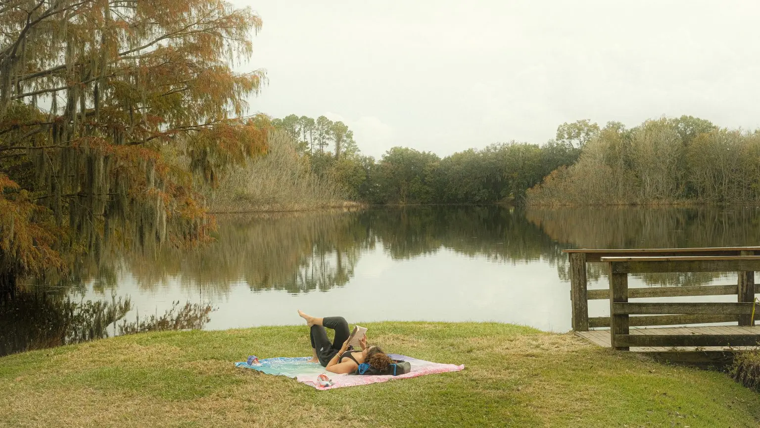 A woman in sports clothes lays on a towel and reads a book on a grassy edge of a lake. She is next to a wooden foot bridge. The foliage is in tones of brown indicating the Fall season.