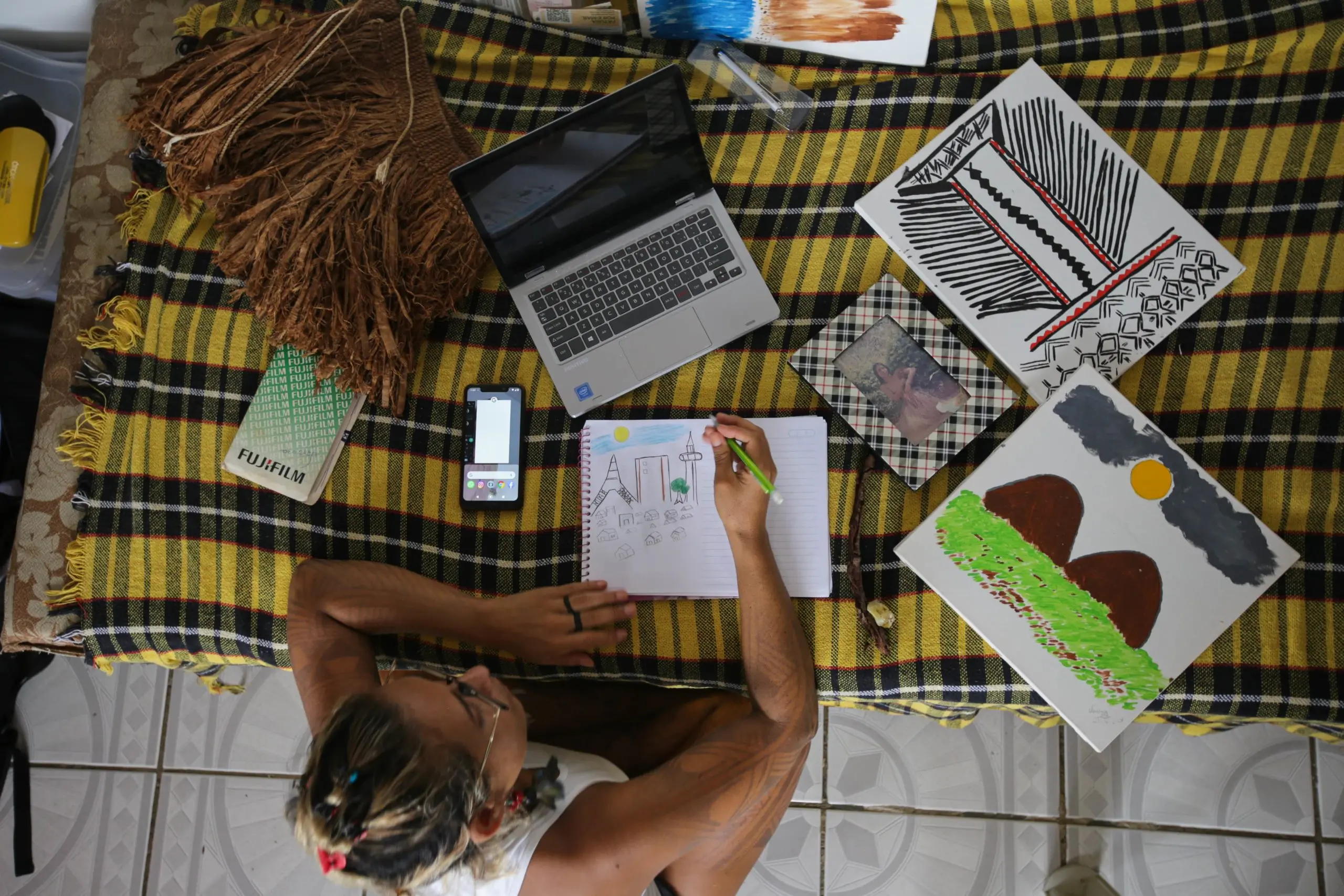 A person works in a notebook, surrounded by a laptop, paintings, a cell phone, and a photo in a frame.