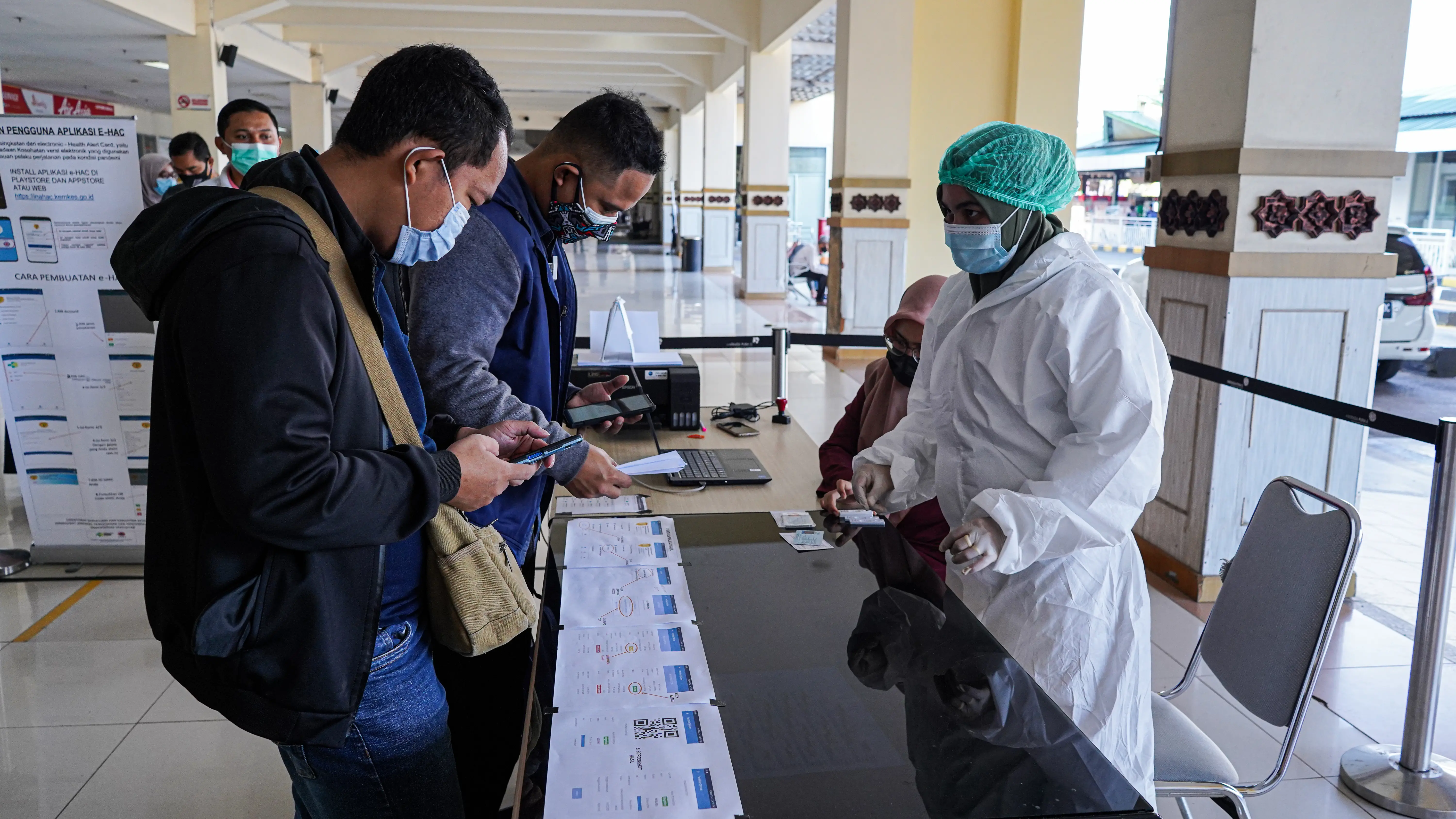 Two men in medical face masks stand at a table across from a person in PPE.