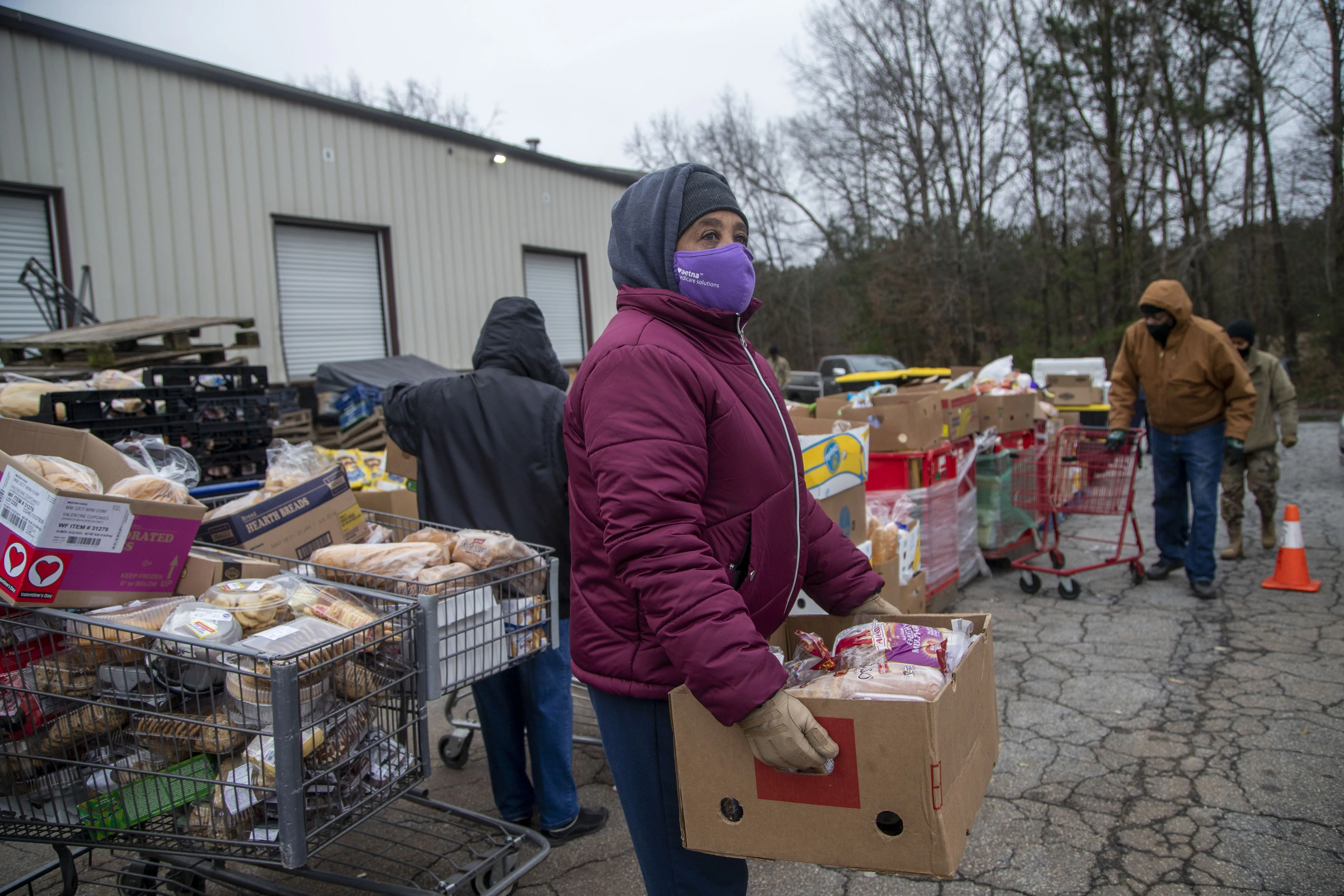A woman wearing a coat and mask holds a box of food.