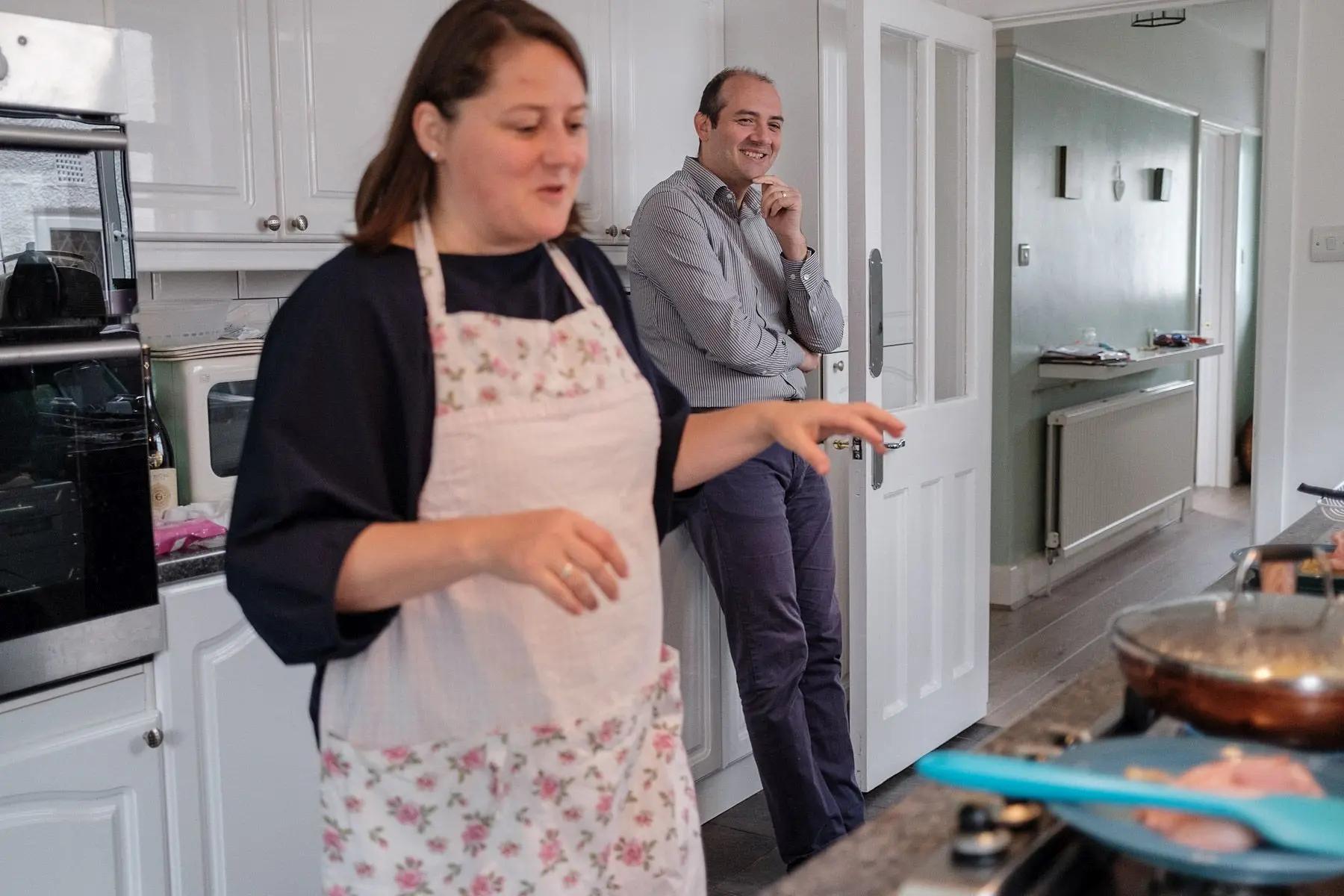 A woman talks and cooks while her husband leans on a kitchen counter, smiling.