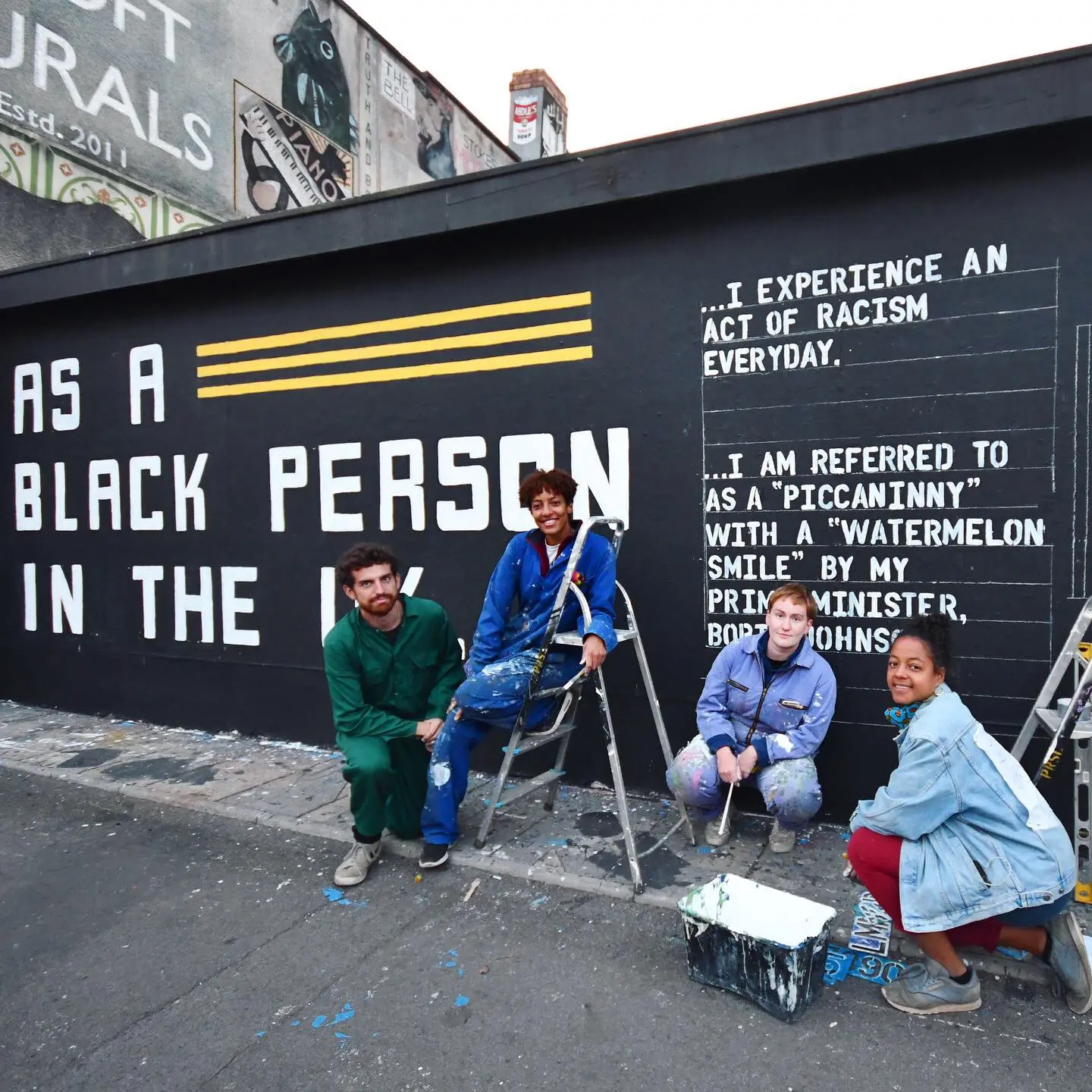 Four people sit in front of a mural that reads, "As a Black person in the U.K."