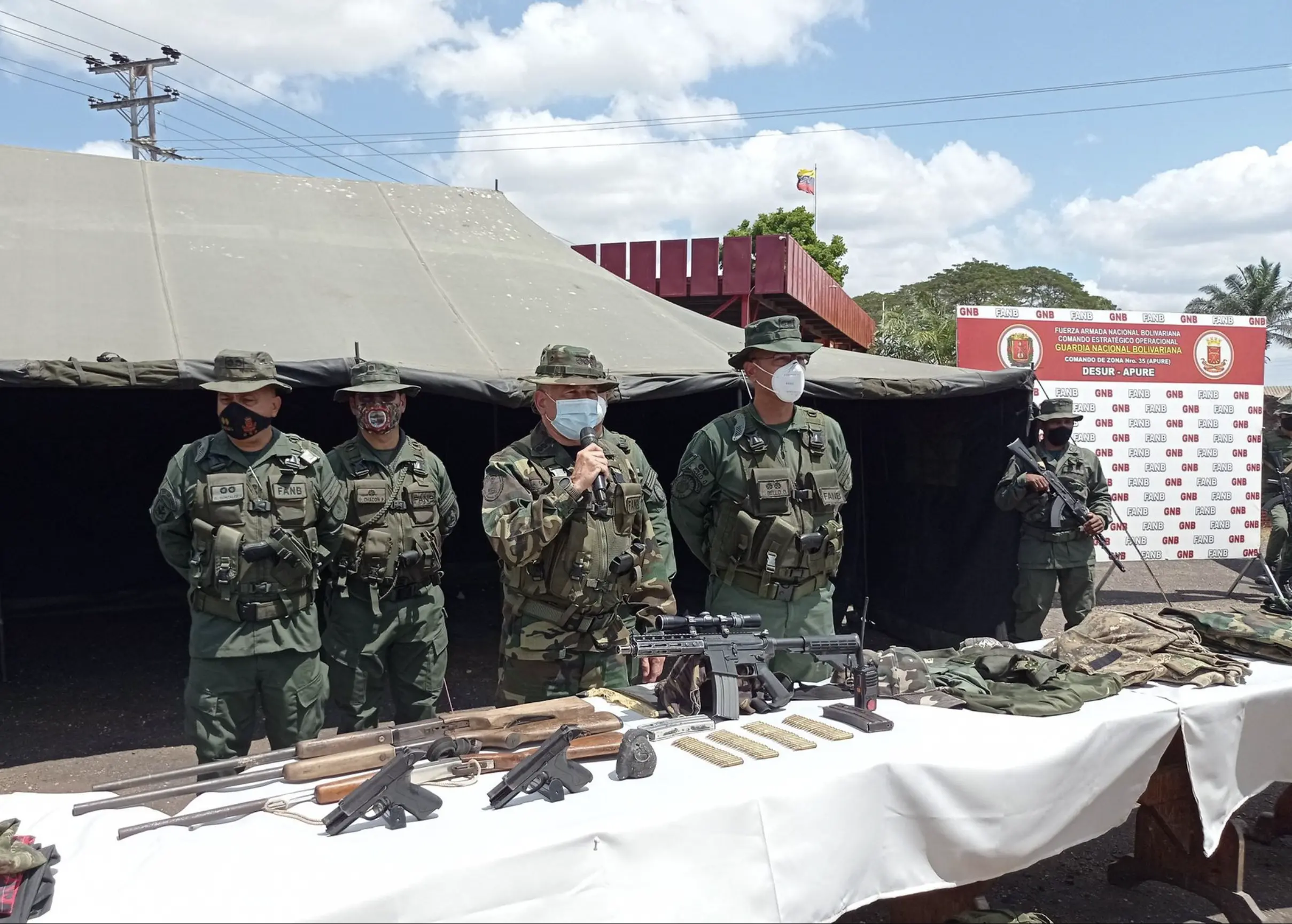 five uniformed men stand behind a table set up in front of a military-style tent. On the table, there are rifles, pistols, bullets, and green military uniforms. In the background, there is a backdrop for photos that reads, in Spanish, "National Bolivarian Armed Forces, Strategic Operational Command, Bolivarian National Guard. Command of Zone number 35 (Apure). DESUR - APURE"