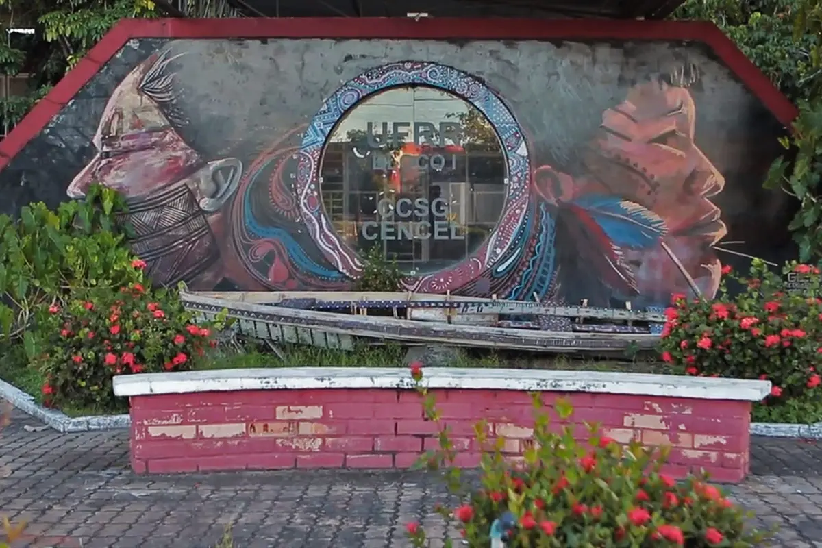 A photo of the entrance to the center of communication, letters, and visual arts at the Federal University of Roraima. Shows mural of Indigenous people and a small brick bench.