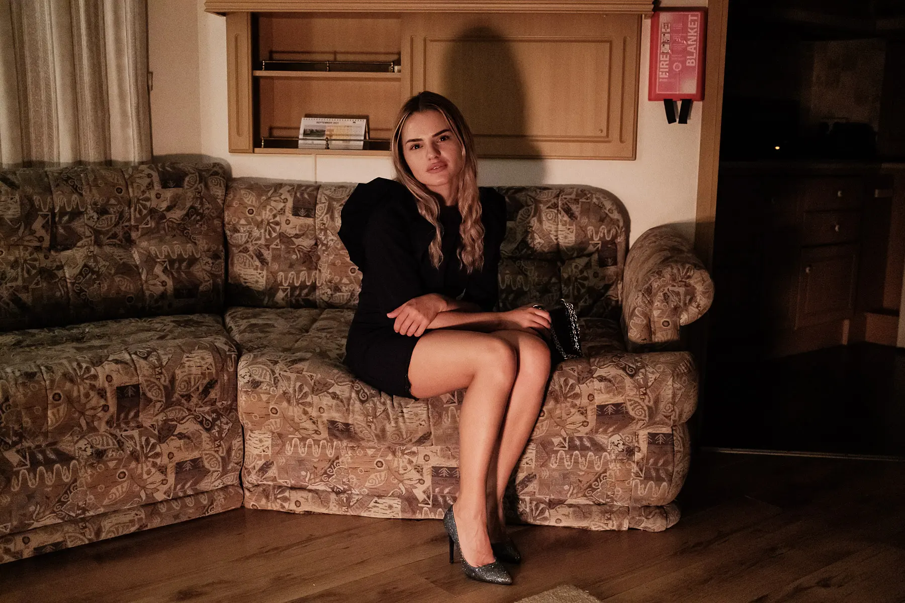 Young woman sits on a couch in her camper in a black party dress and heels 