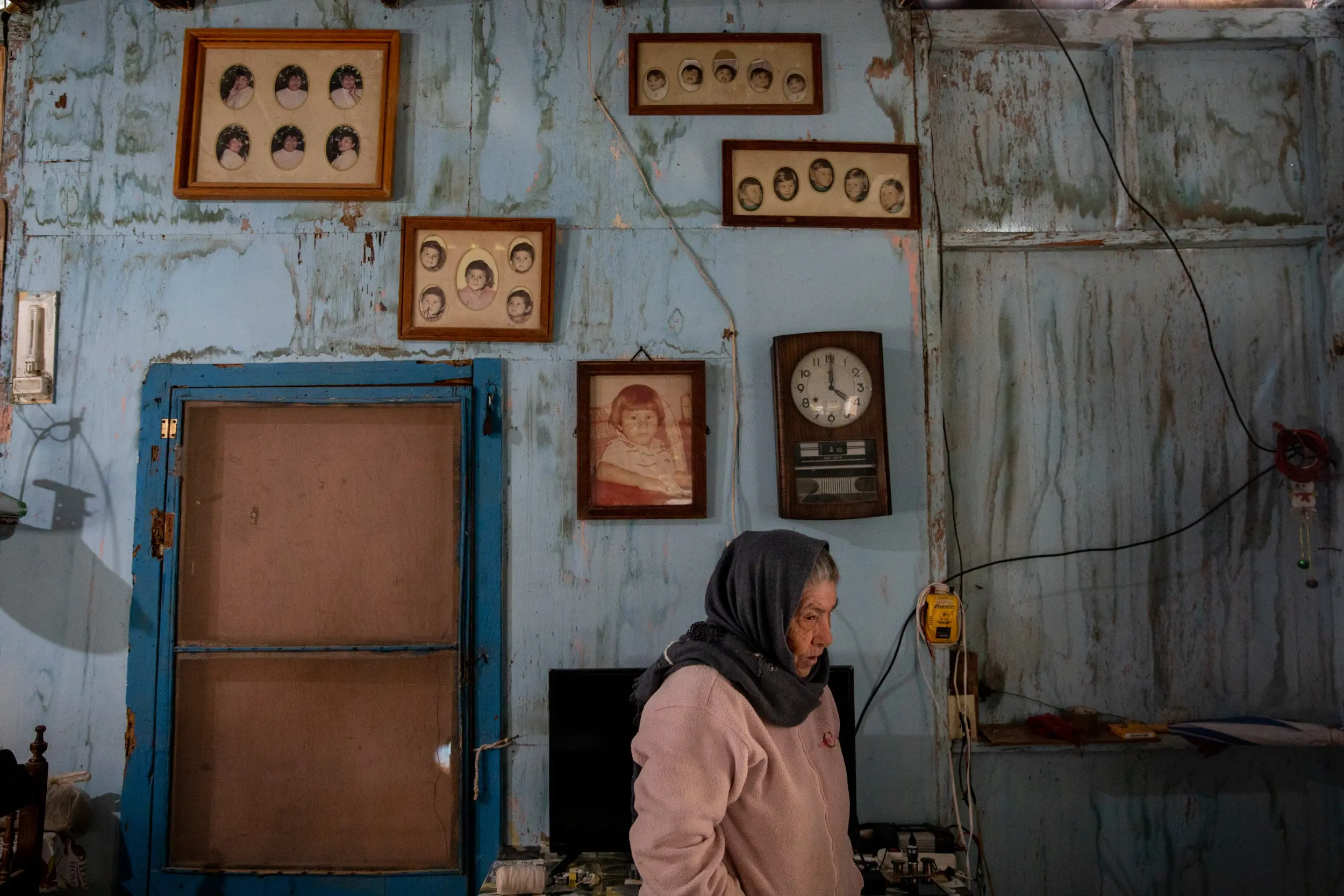 A woman stands in front of a wall with childhood photos, and a clock.