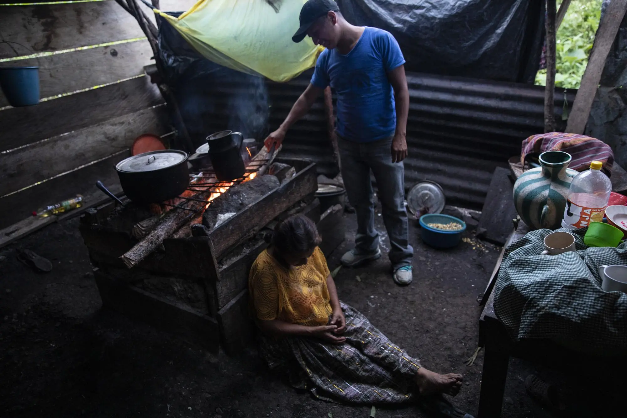 A woman sits on ground in front of a stove while her sons cooks