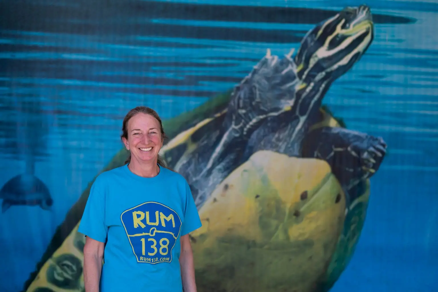 a woman smiles for a picture in front of a large print of a freshwater turtle. Her Shirt reads Rum 138 rum138.com