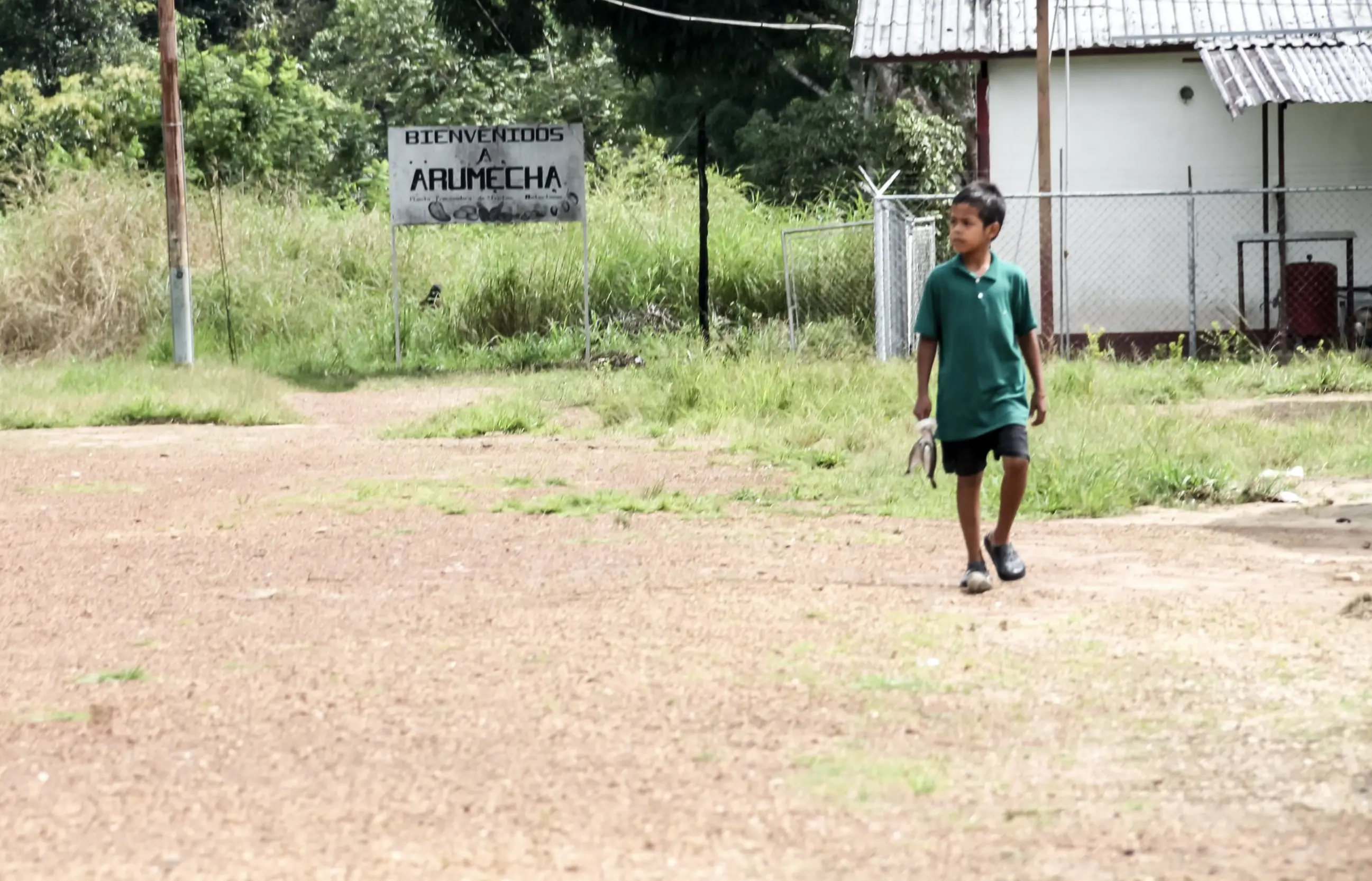 A young boy walks along a path with a Zinc metal roofed building behind him. In the background, a sign reads "Welcome to Arumecha" in Spanish.