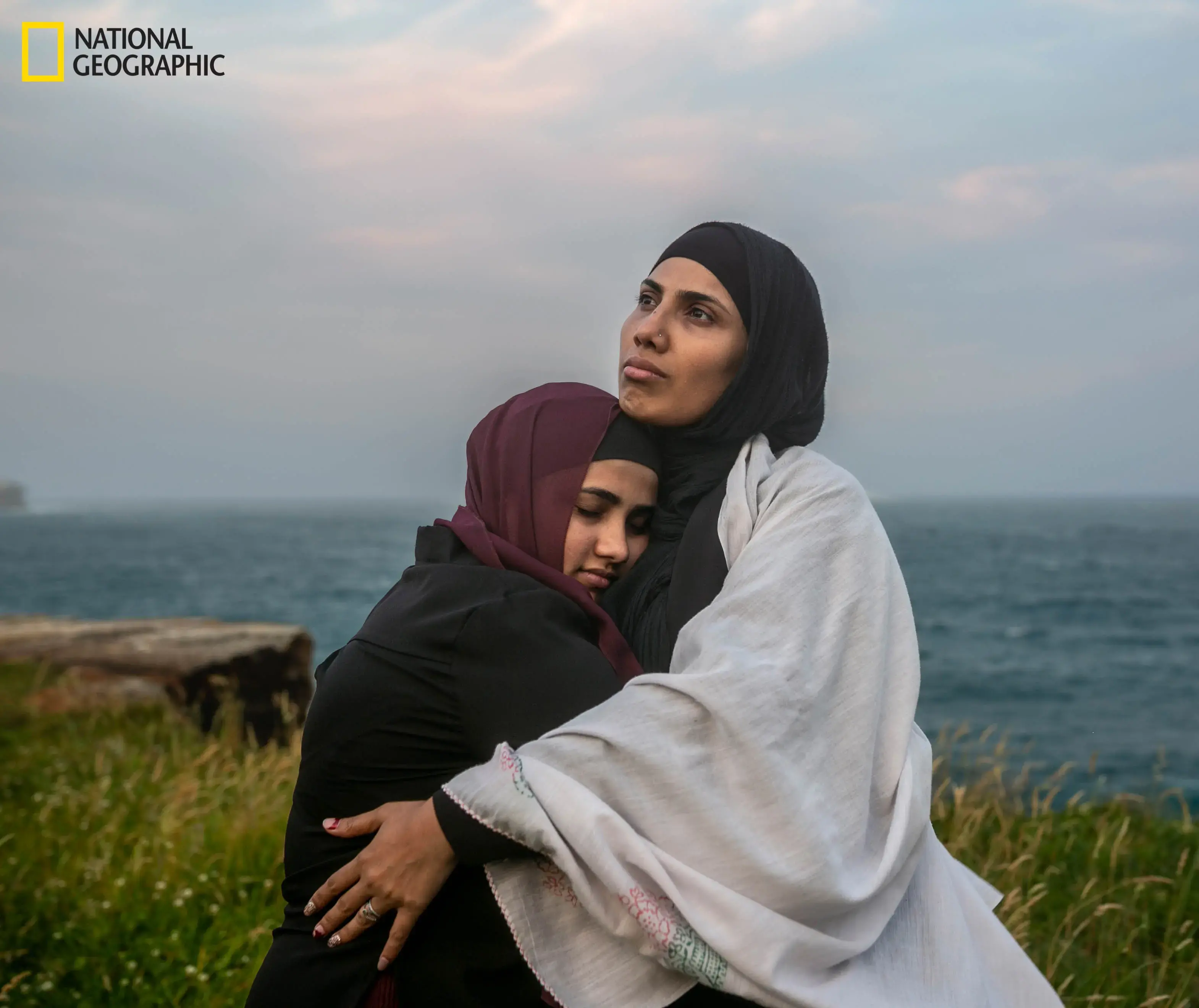 Sajeda Bahadurmia, 32, embraces the eldest of her six children, Asma, 16, on a beach near Sydney. Sajeda is wrapped in a white scarf her mother gave her as a safety charm before their gruel- ing three-month jour- ney six years before. As Rohingya Muslims, they fled persecution in Myanmar. But the haven they found in Australia is an uncer- tain one because they don’t yet have perma- nent residency. Image by Mridula Amin. Australia, 2020. Media file: mm9019_ngs-618315-19_191102_07741.jpg